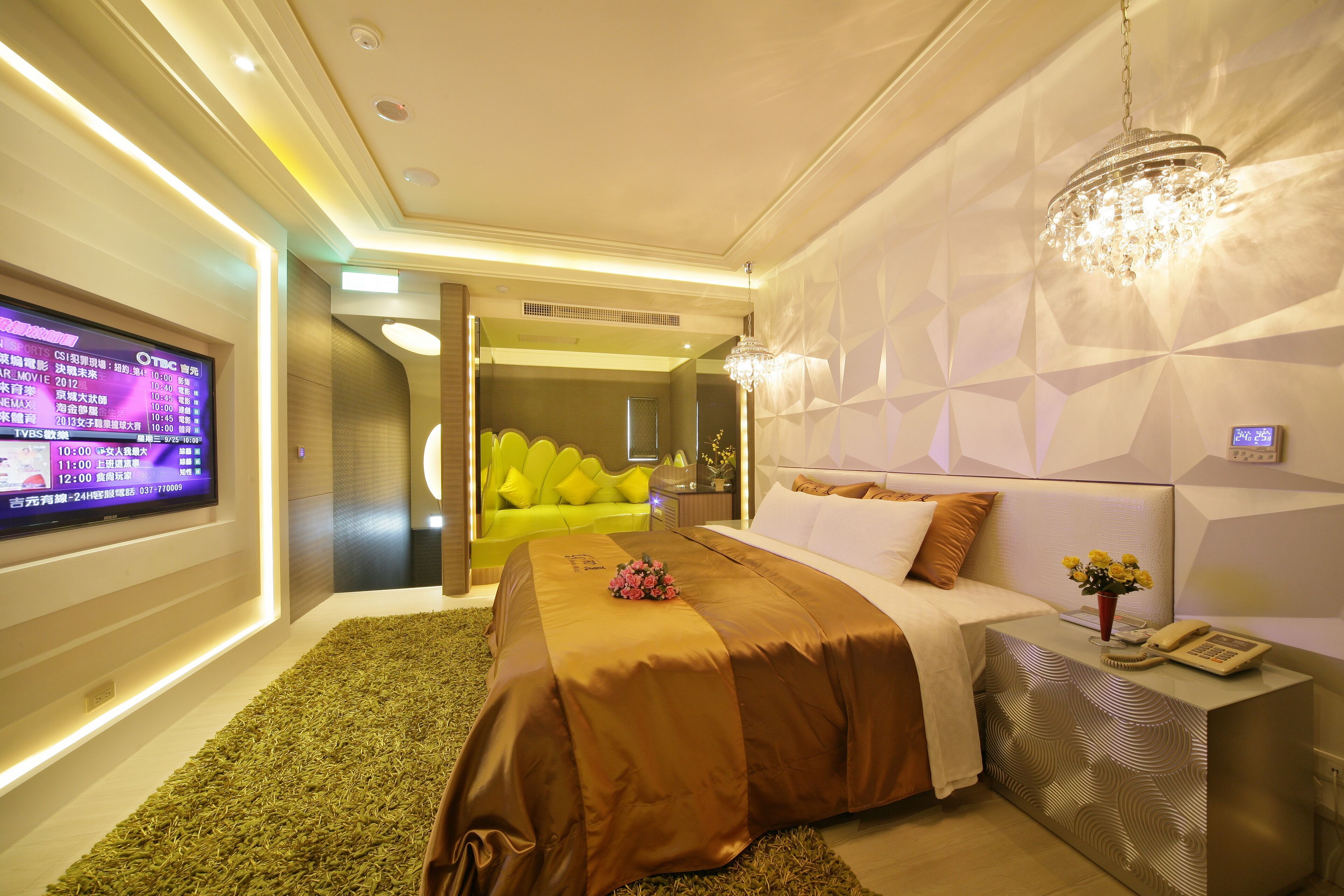 Una habitación de hotel moderna con un televisor de pantalla plana grande, un sofá verde, una cama con colcha marrón y un candelabro decorativo.