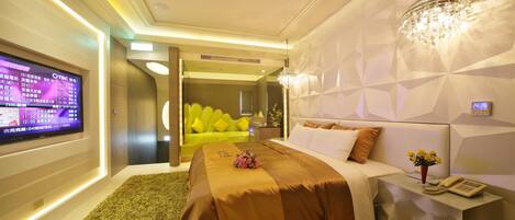 Una habitación de hotel moderna con un televisor de pantalla plana grande, un sofá verde, una cama con colcha marrón y un candelabro decorativo.