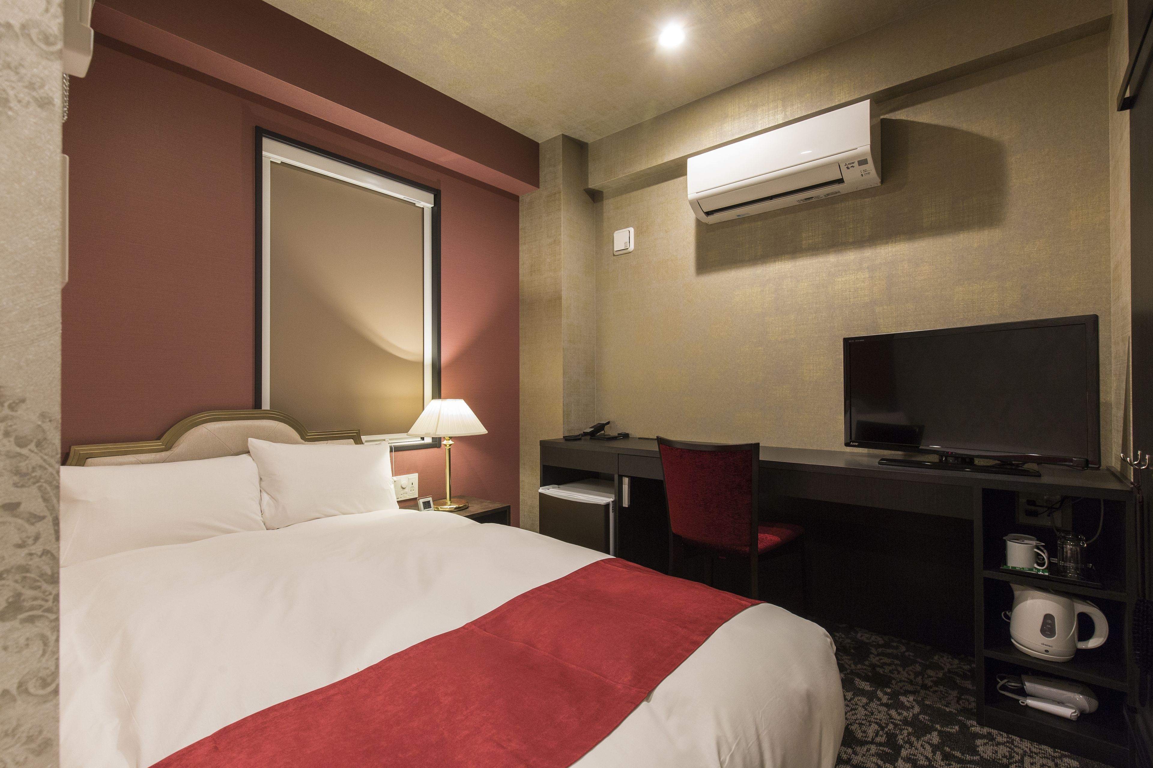 Habitación de hotel con cama, escritorio, silla, televisor y estante con tetera y otros objetos.