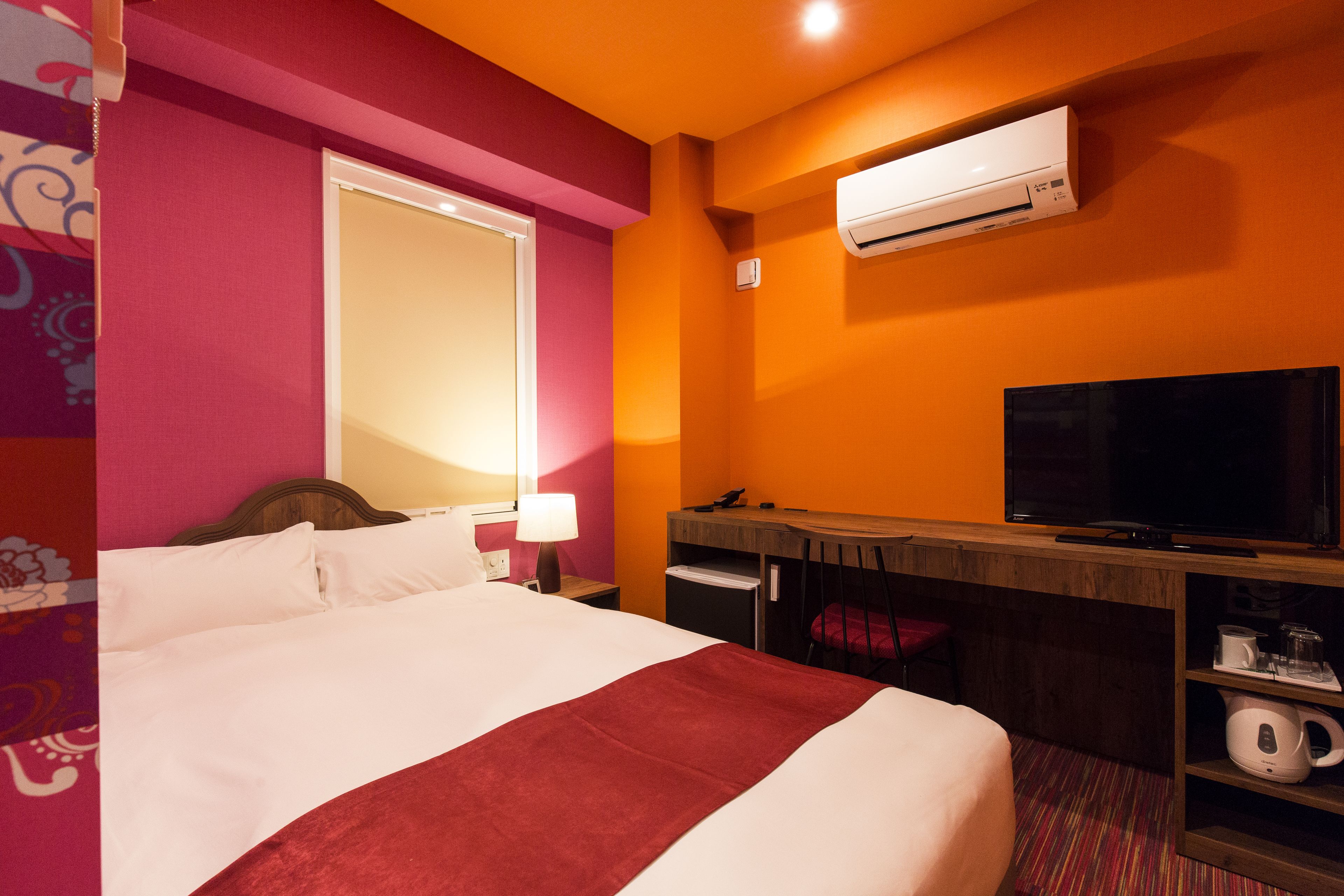 A hotel room with a bed, a desk with a television, an air conditioner, and a small kitchenette.