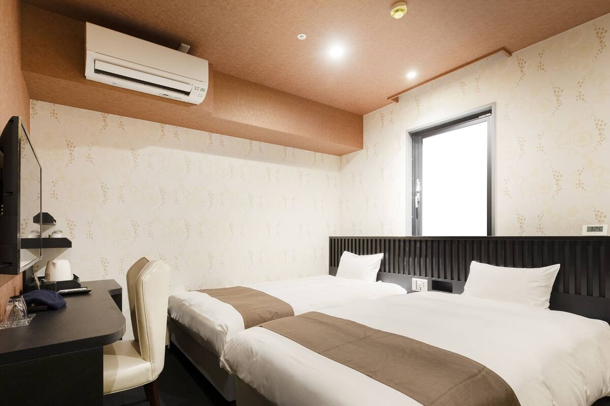 Habitación de hotel con dos camas, un escritorio con silla, aire acondicionado y ventana con vistas.