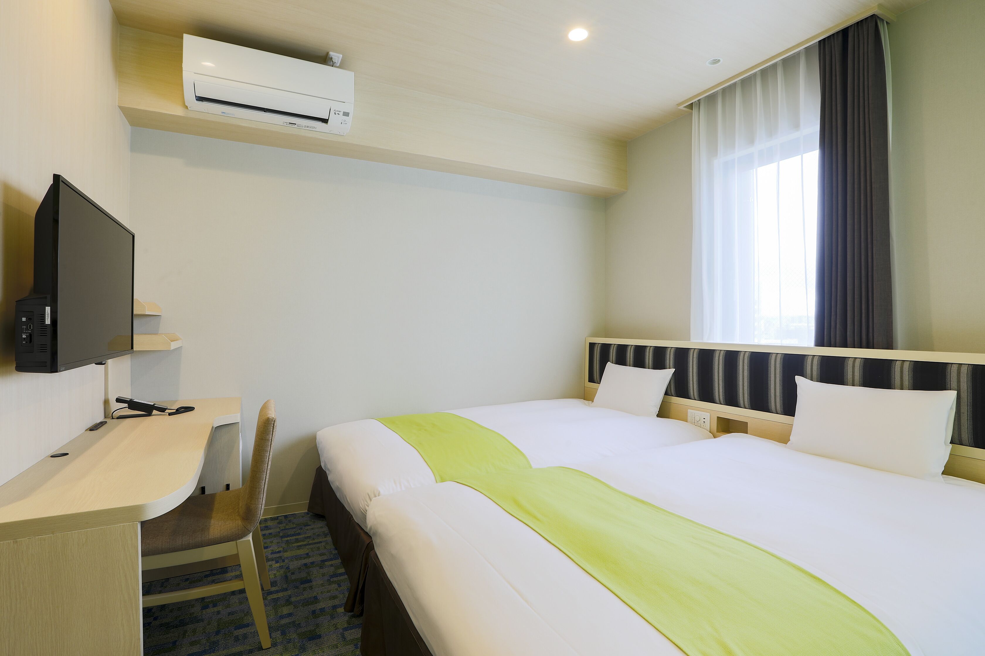 A hotel room with a bed, a desk, a chair, a television, and an air conditioning unit.
