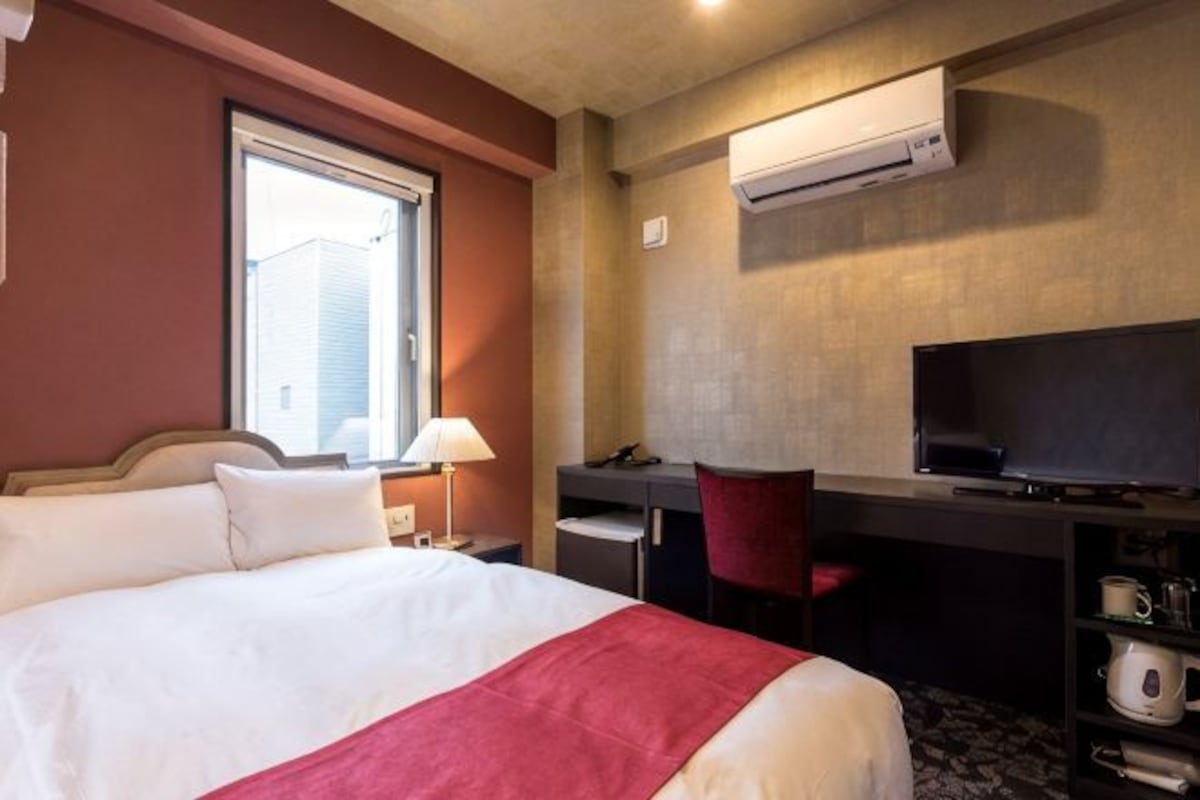 A hotel room with a bed, desk, chair, TV, and an air conditioning unit.