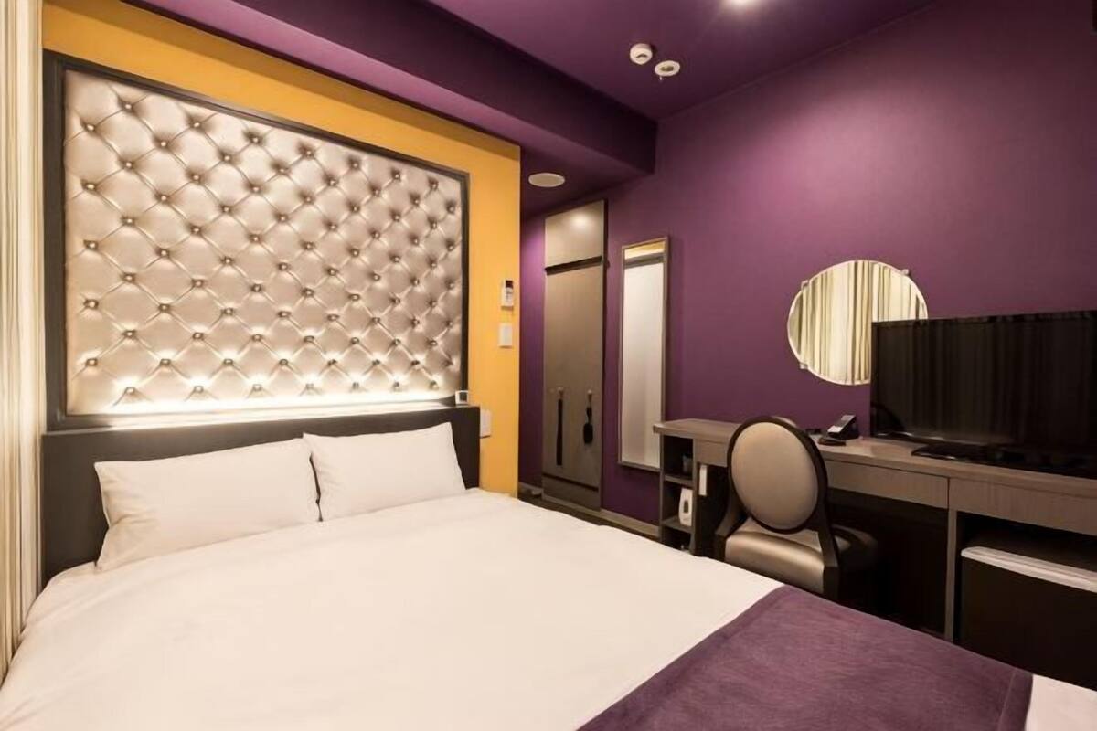A hotel room with a tufted headboard, a bed, a desk with a chair, a television, and a mirror.