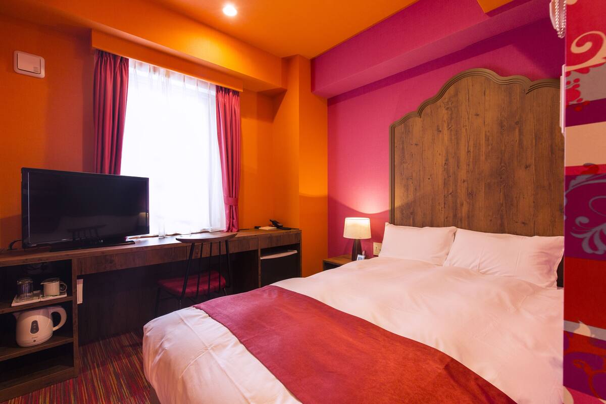 Habitación de hotel con cama, escritorio con televisor, una silla, mesita de noche con lámpara y ventana con cortinas.