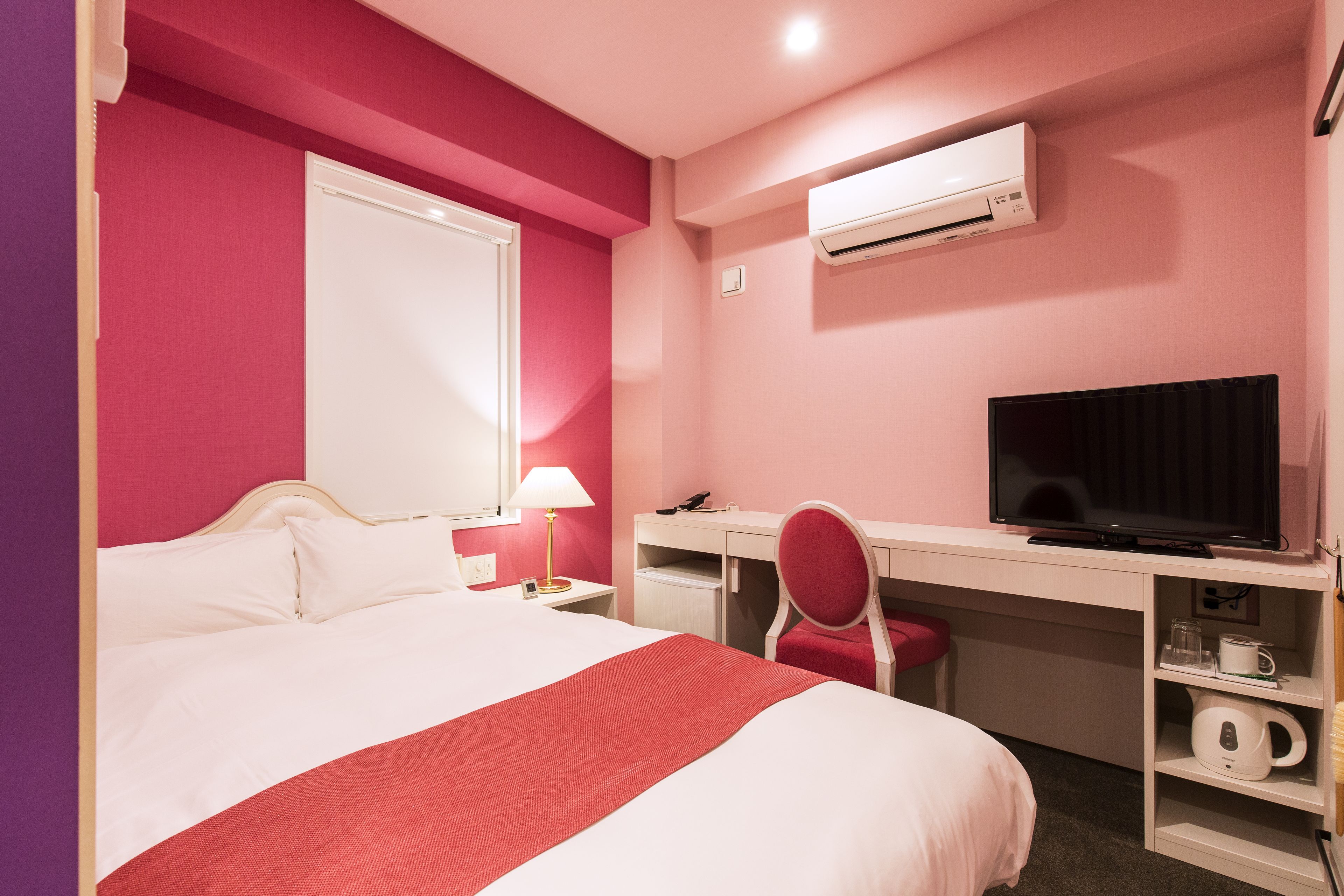 A hotel room with a bed, a desk, a chair, a television, and an air conditioning unit.