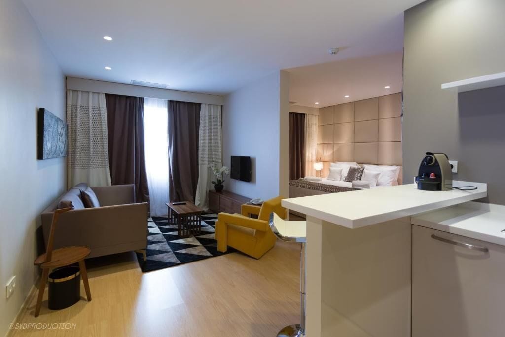 A modern hotel room with a sofa, dining table, yellow chairs, a kitchen area, and a bed.