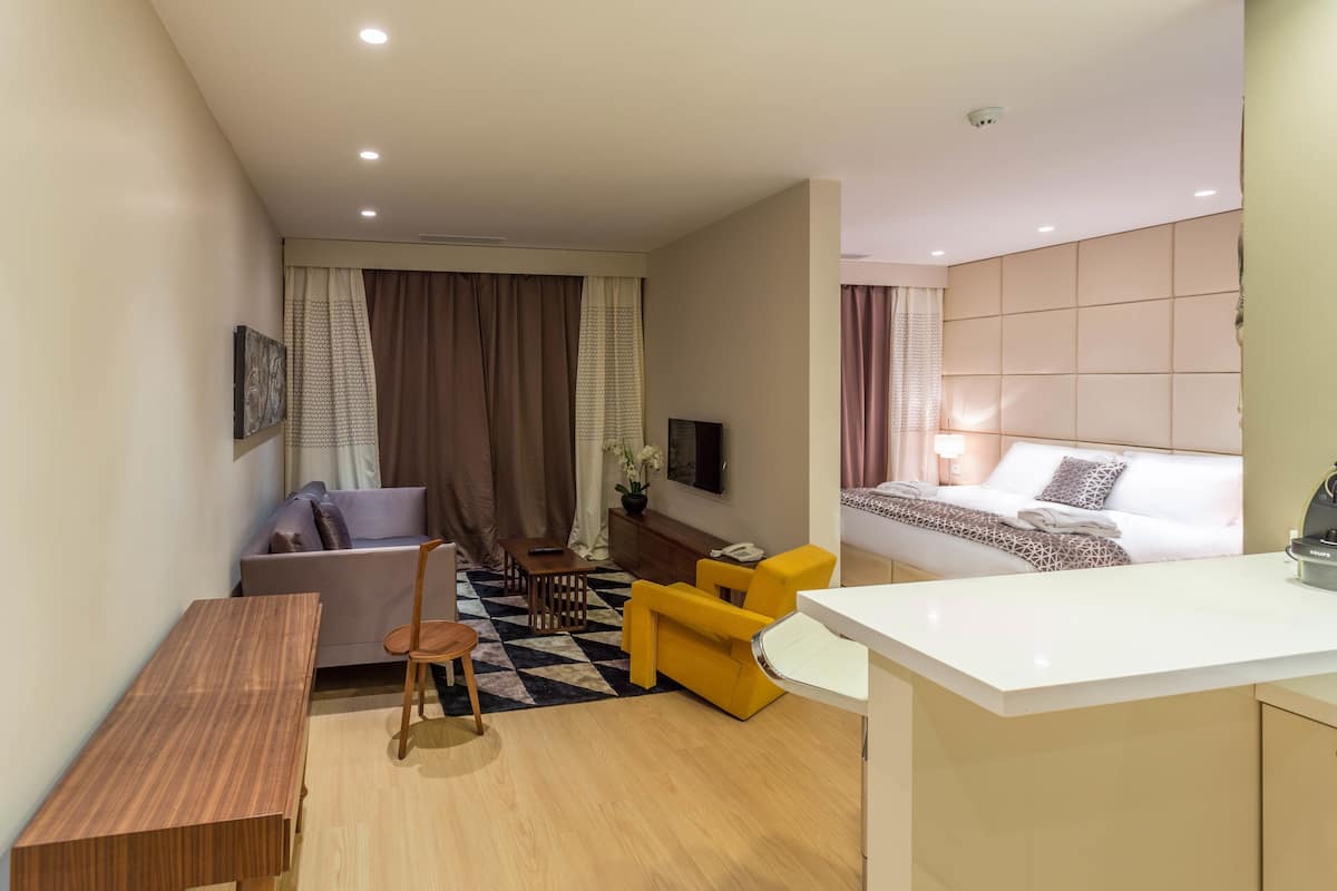 A modern hotel room with a bed, a sofa, a dining table, and a small kitchen area.