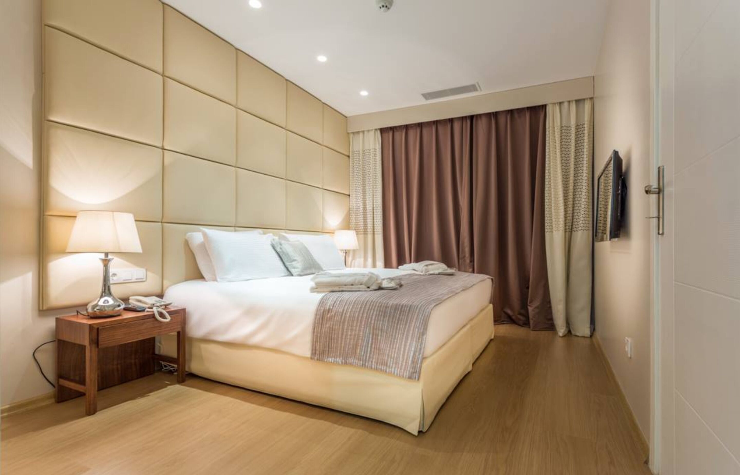 A hotel room with a large bed, a bedside table with a lamp and phone, and a view of the outside through curtains.