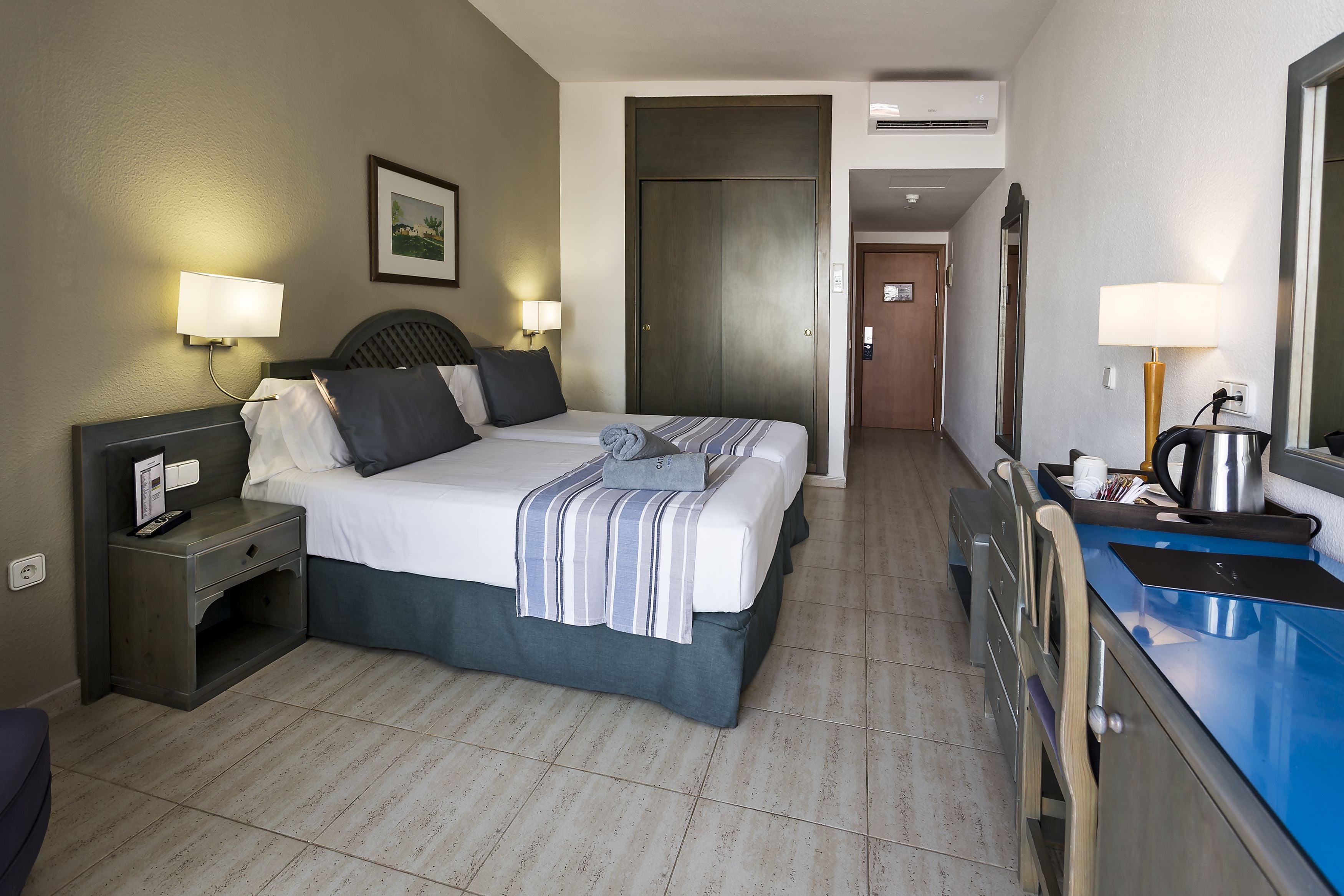 A hotel room with a bed, bedside lamps, a desk with a lamp, a mirror, and a bathroom counter with a sink and a kettle.
