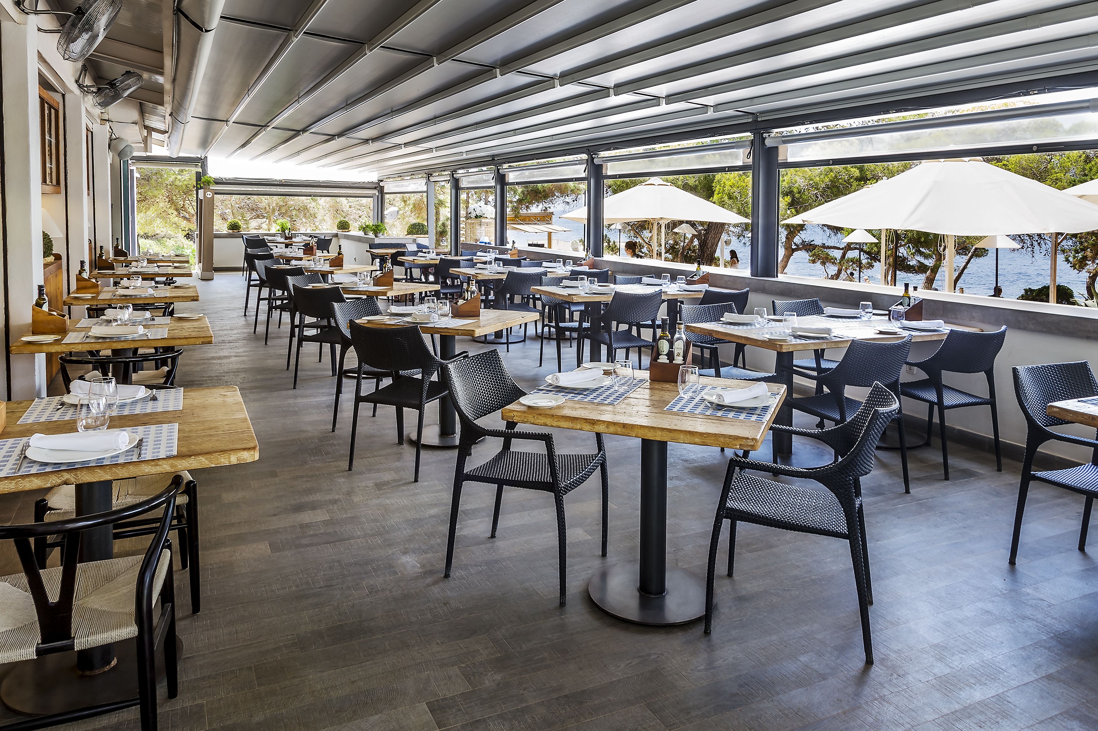 Un área de comedor al aire libre con mesas y sillas, grandes sombrillas y vista al mar.