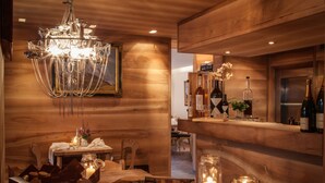 A wooden interior with a chandelier, a dining table with candles, and a bar area with bottles and glasses. - Restaurant - Hotel Sonnmatten (Zermatt)