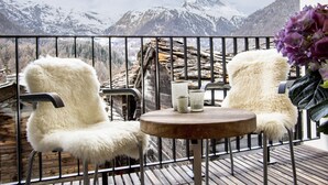 A balcony with two white furry chairs, a round wooden table, and a vase of purple flowers. - Restaurant - Hotel Sonnmatten (Zermatt)