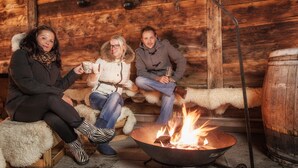 Three people sitting by a fire in a rustic cabin with wooden walls and a barrel. - Restaurant - Hotel Sonnmatten (Zermatt)