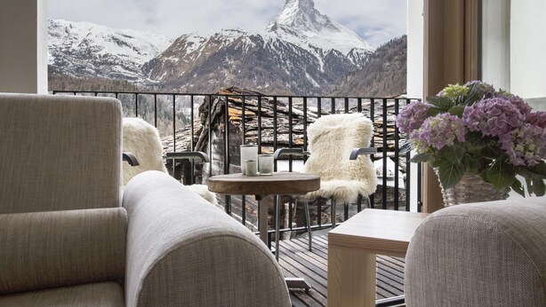 A balcony with a view of snow-capped mountains, a wooden table, and two chairs with white fur throws. - Restaurant - Hotel Sonnmatten (Zermatt)