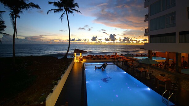 A pool area with a view of the ocean at sunset, palm trees, and outdoor seating. - Sevonra Garden (Unawatuna)