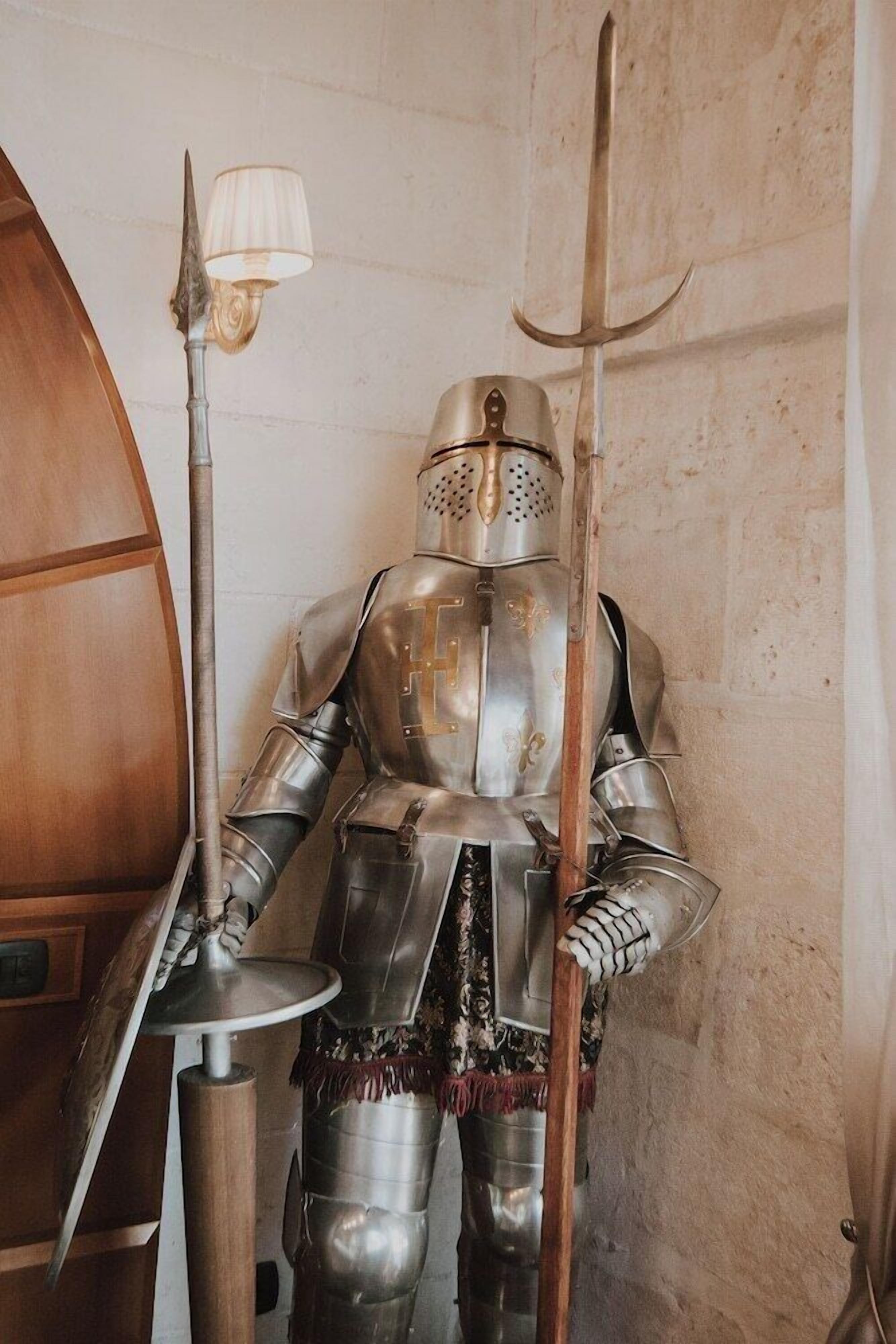 Un caballero medieval con armadura completa de pie en una habitación con una silla de madera y una lámpara de mesa.