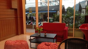 Une pièce avec un canapé rouge, deux poufs à motifs, une petite table avec une plante, et une vue sur une rue bordée de voitures garées et d’immeubles.