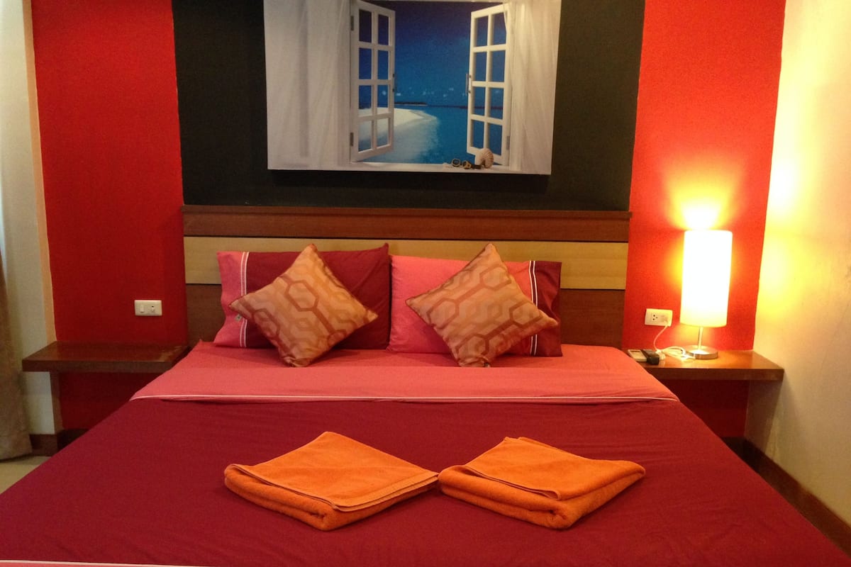A hotel room with a large bed, red walls, a painting of an open window, and a night light on the nightstand.
