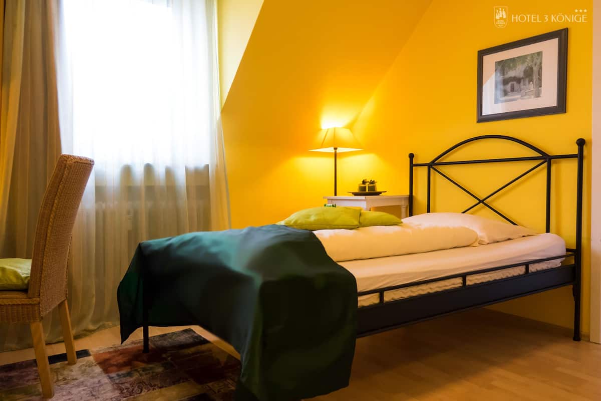 Una cama individual con cubrecama verde, una mesita de noche con lámpara, una silla y un cuadro en la pared.