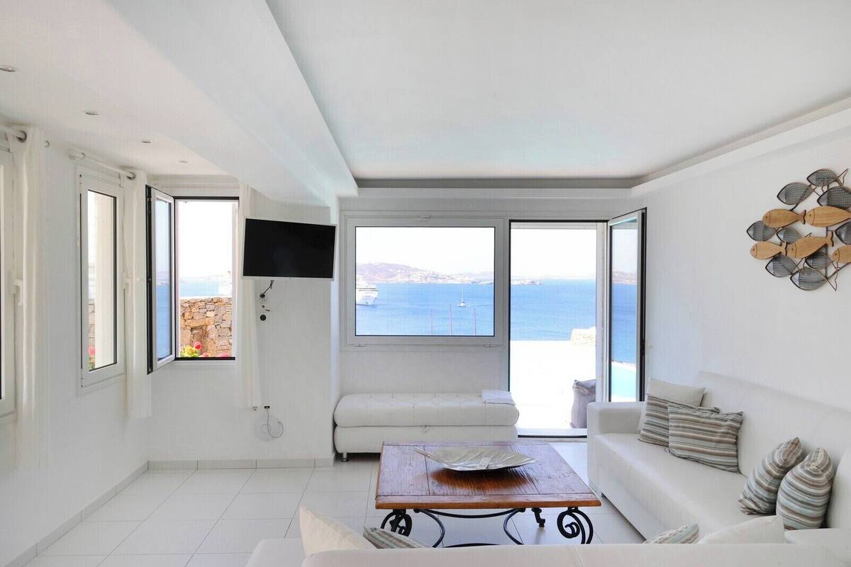 A spacious living room with a large window overlooking the sea, a white sofa, a coffee table, and a wall-mounted TV.