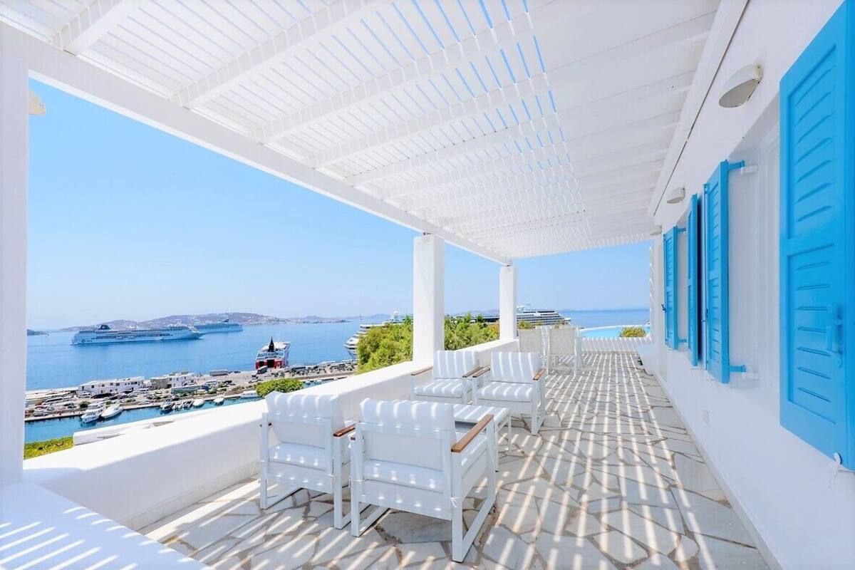 A covered outdoor seating area with white chairs and blue shutters, overlooking a marina and the sea.