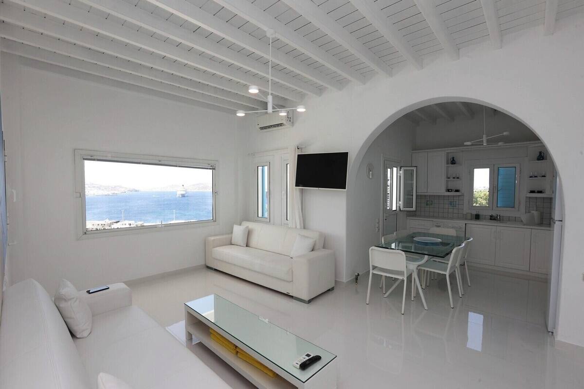 A modern living room with a white sofa, a glass coffee table, and a dining area with a glass table and chairs. There is a TV mounted on the wall and a view of the sea through a large window.