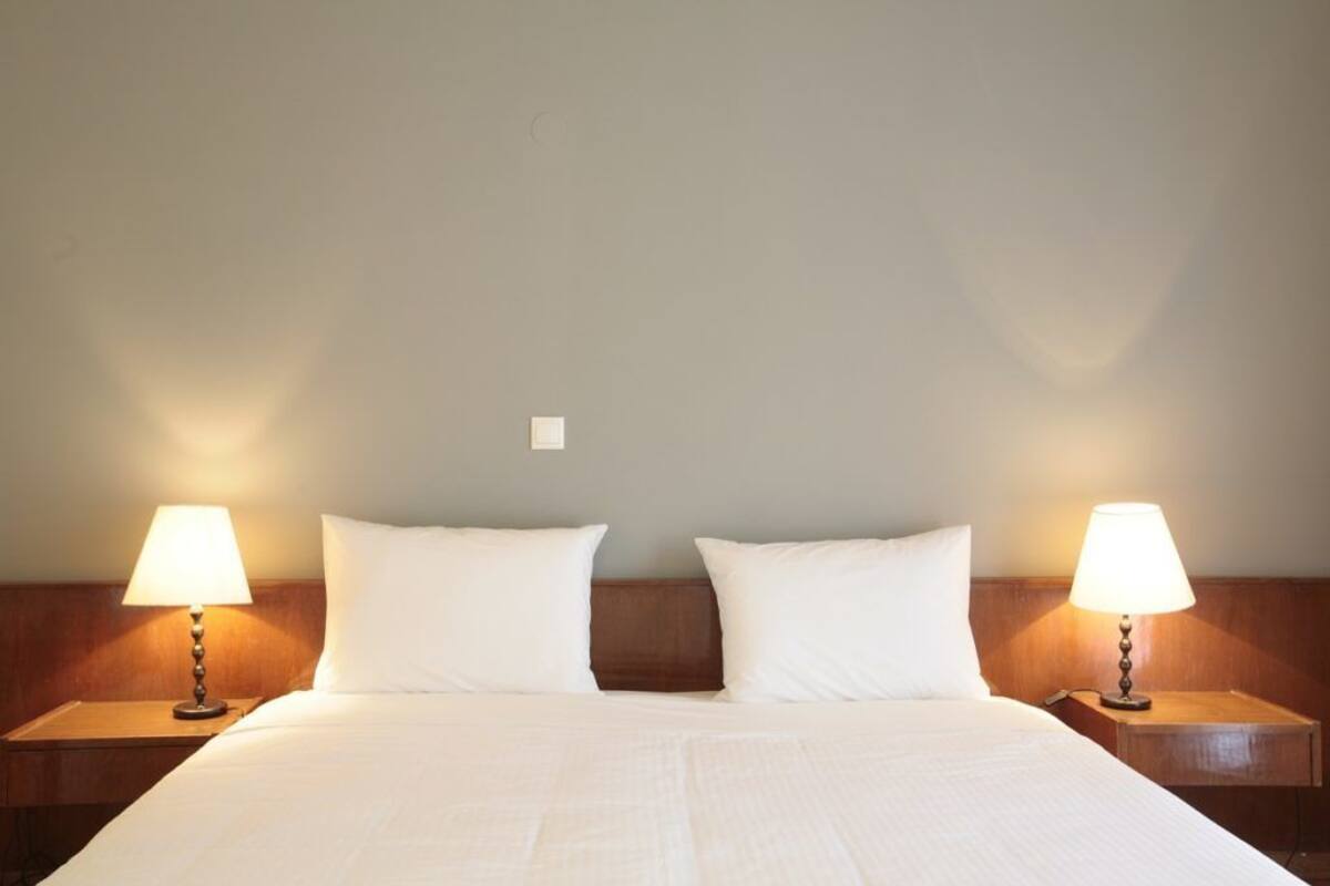 Una cama bien hecha con sábanas blancas, flanqueada por dos mesitas de noche con lámparas, en una habitación sencilla con pared lisa y cabecera de madera.