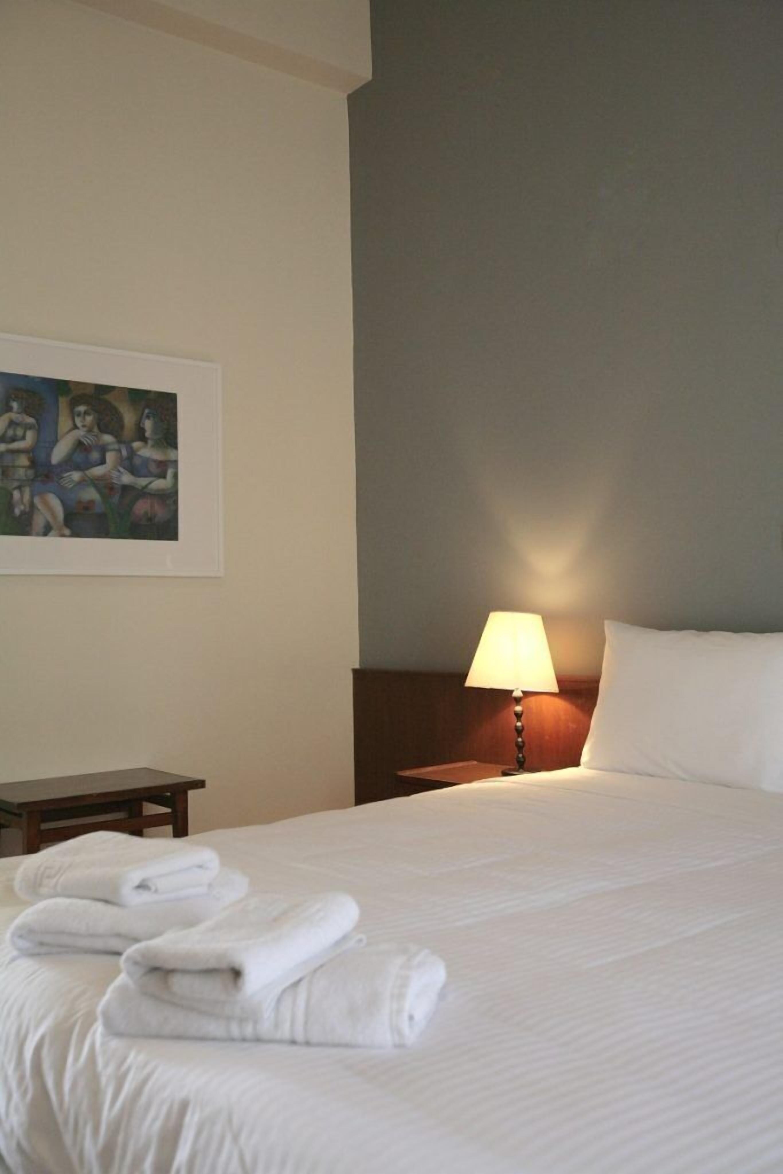 Habitación de hotel con una cama bien hecha, una lámpara de noche, una mesita y un cuadro en la pared.