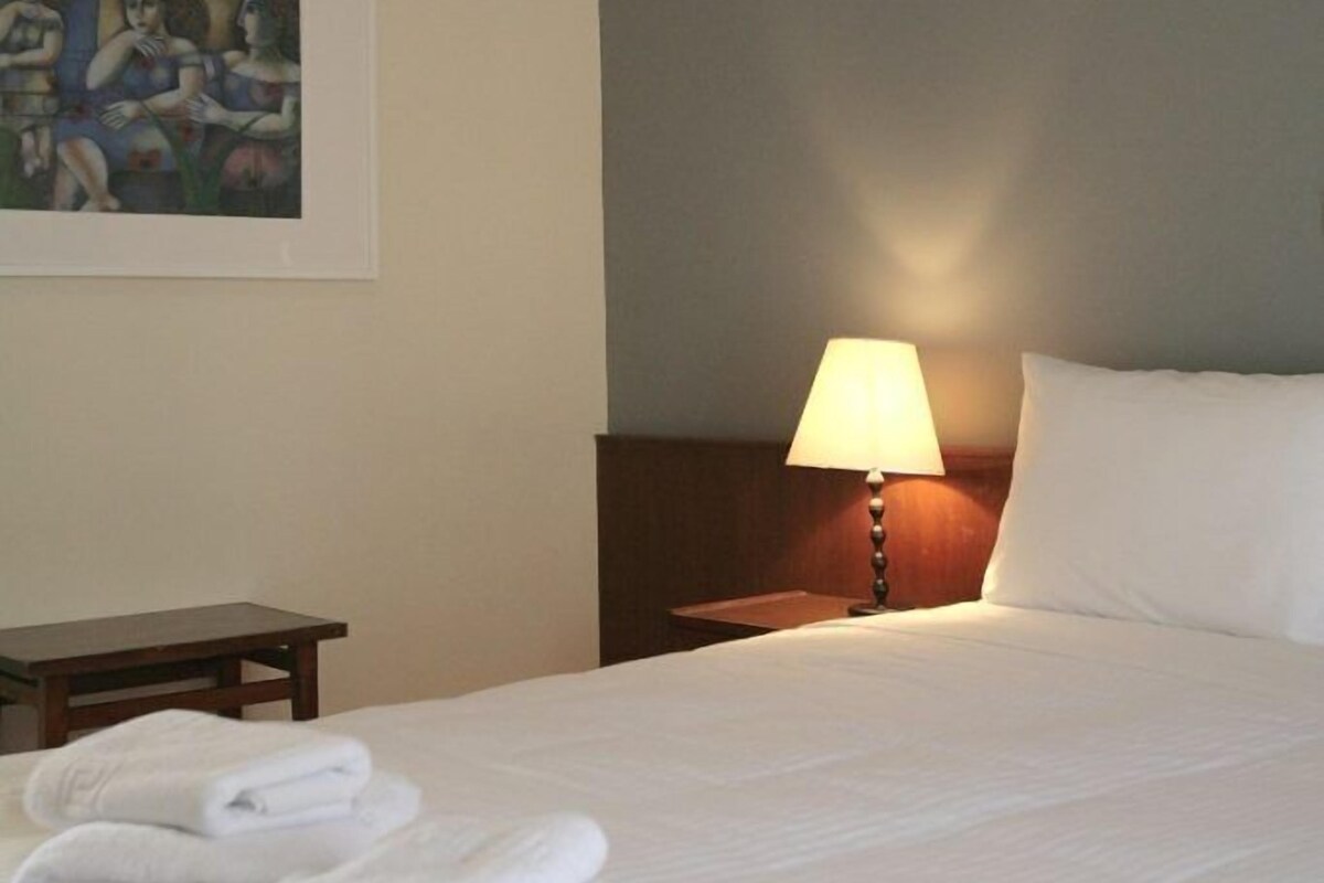 Habitación de hotel con una cama bien hecha, una lámpara de noche, una mesita y un cuadro en la pared.