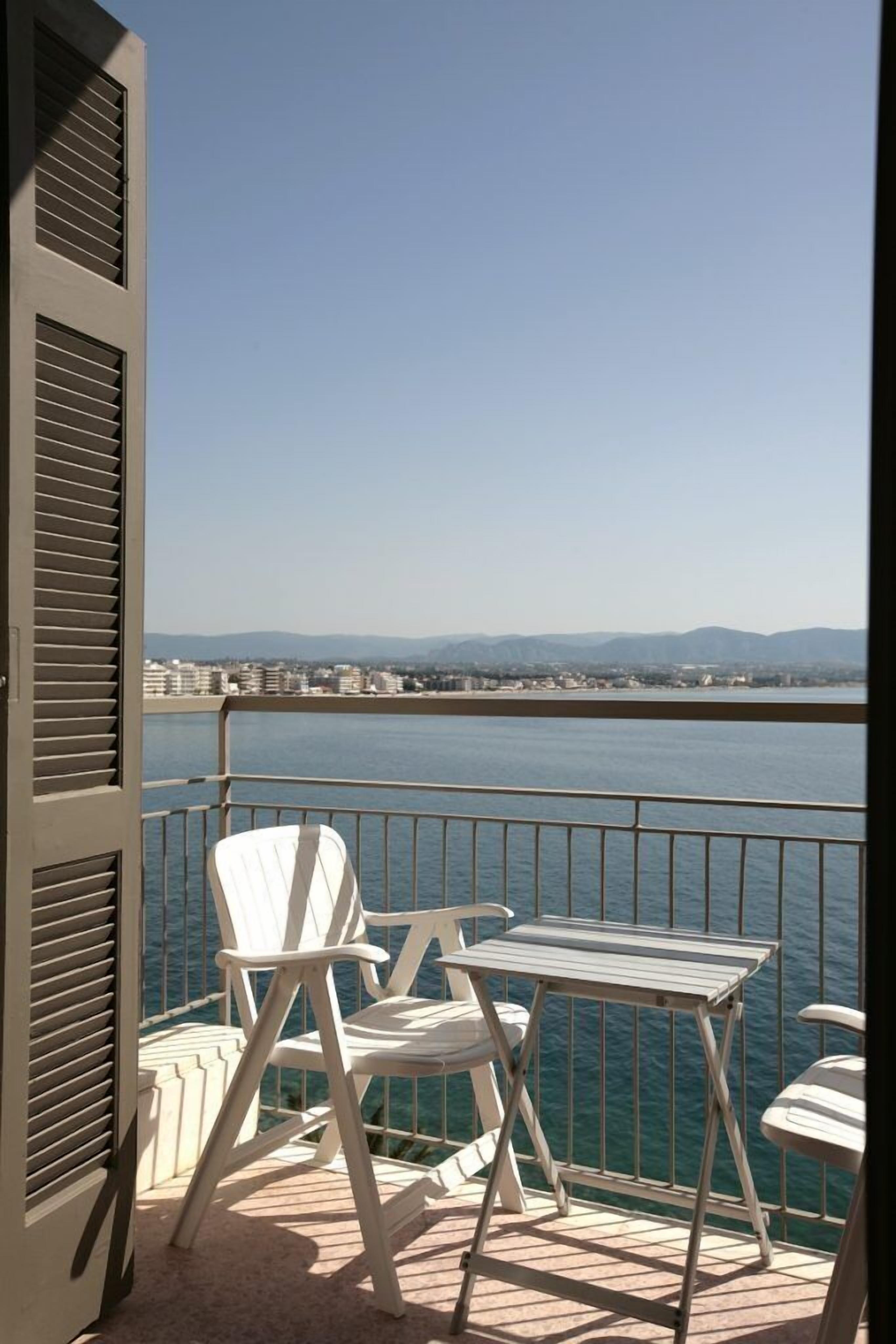 A balcony with two white chairs and a small table, overlooking a body of water and distant cityscape.