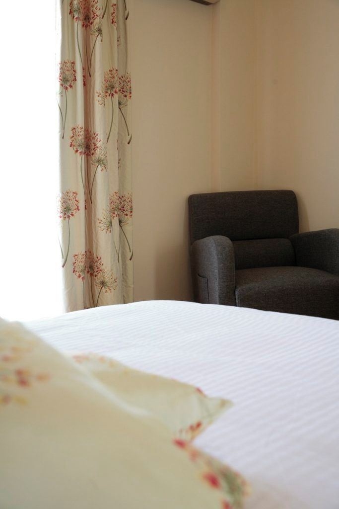 Quarto de hotel com uma cama, uma poltrona cinzenta e cortina com padrão floral.