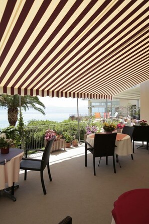 An outdoor dining area with striped awnings, tables, chairs, potted plants, and a view of the sea. - Hotel Theoxenia (Loutraki-Agioi Theodoroi)