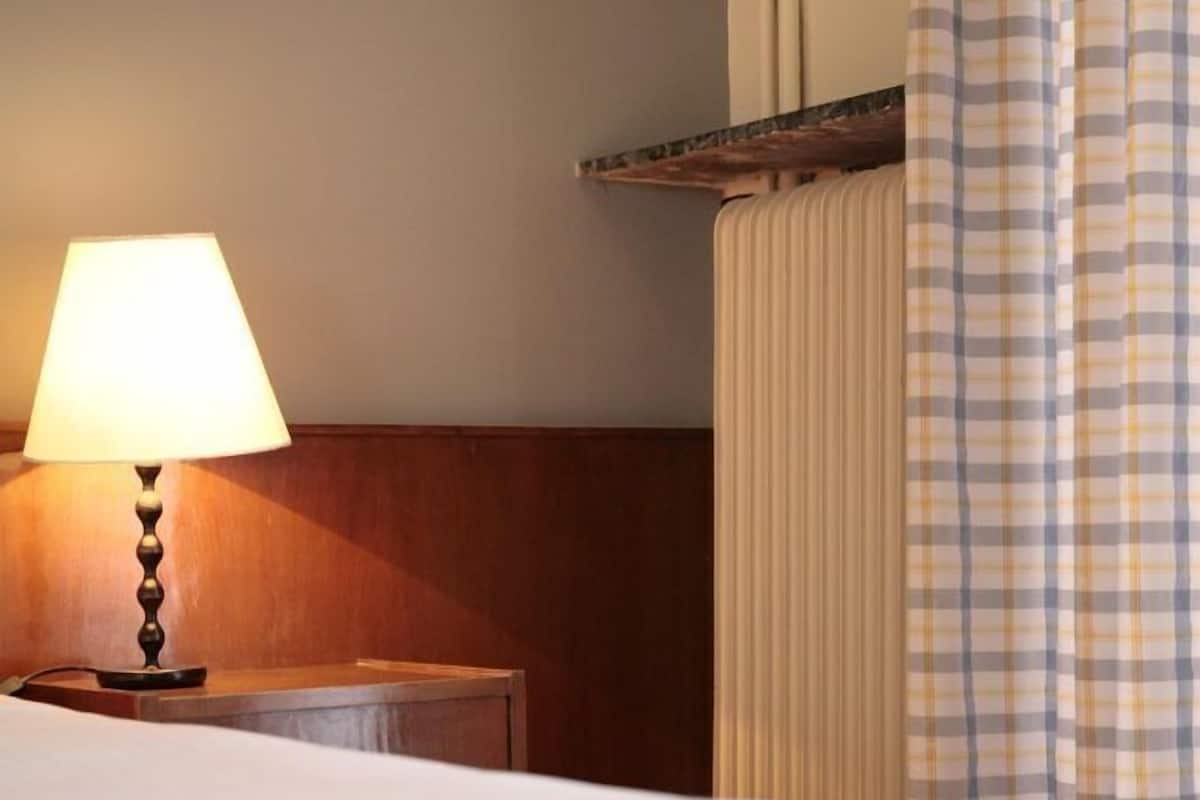 Una habitación de hotel con una cama, una lámpara de noche, un cabecero de madera y una cortina a cuadros.