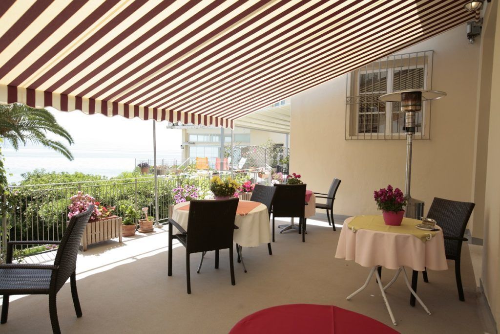An outdoor dining area with striped awnings, tables with chairs, potted plants, and a view of the sea.