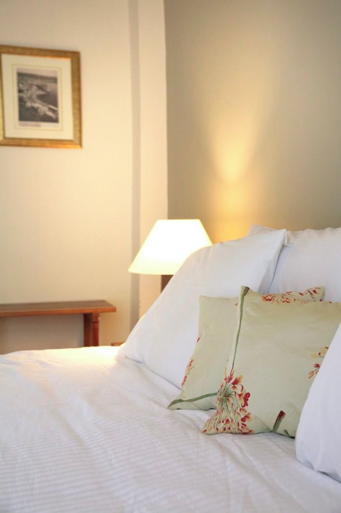 Un lit bien fait, avec du linge de lit blanc et des coussins à motifs floraux, une table de chevet avec une lampe et un tableau encadré au mur.