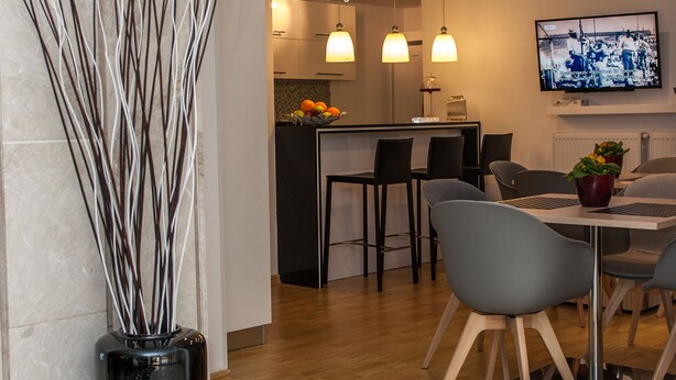 Une cuisine moderne avec un coin repas, agrémentée d’une colonne en marbre, d’un vase noir orné de branches et d’un téléviseur fixé au mur.