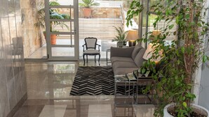 Un salon moderne avec un canapé, un fauteuil et une table basse. Il y a une grande fenêtre avec des stores, une plante en pot et un sol carrelé.
