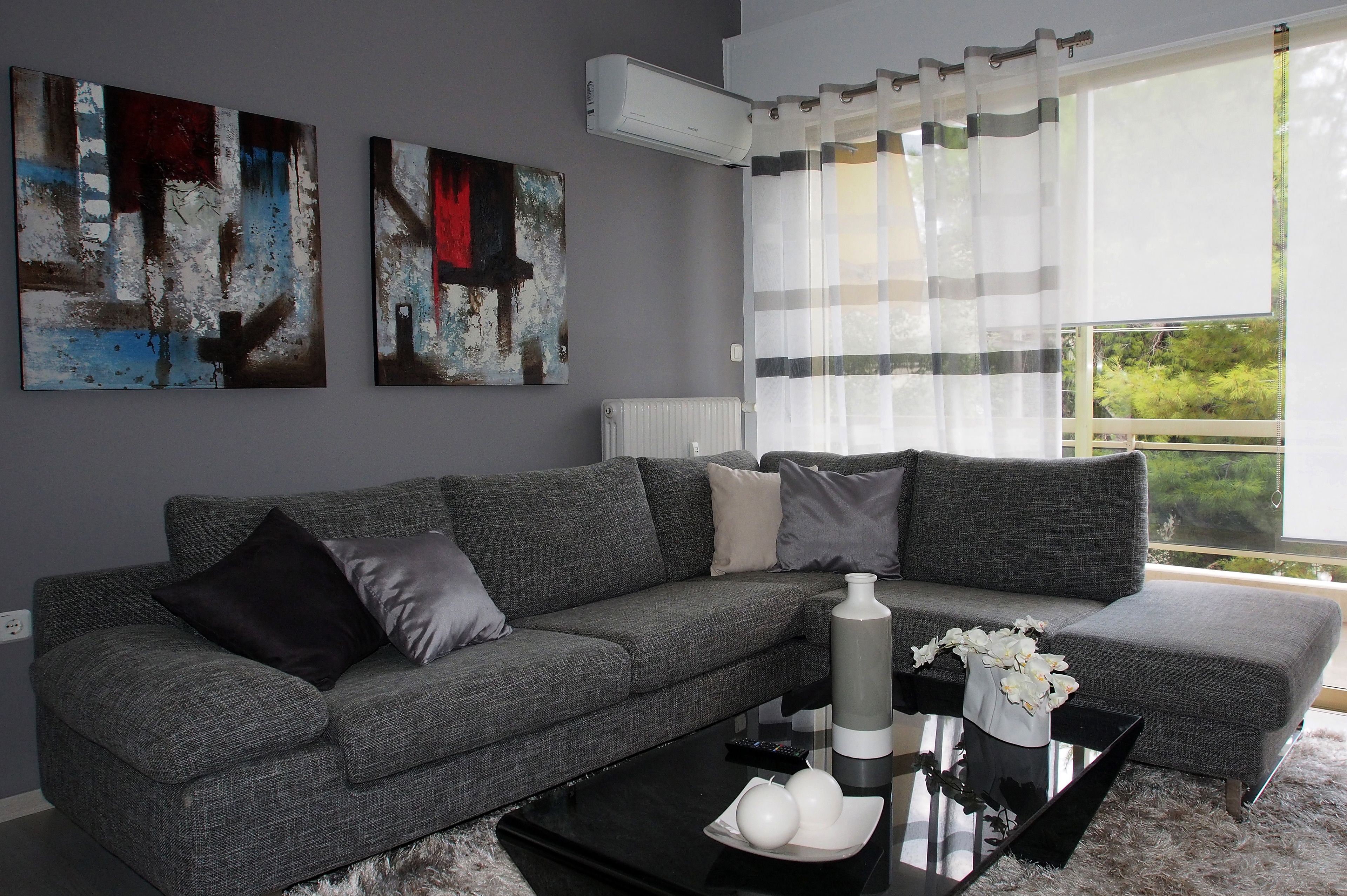 A modern living room with a grey sofa, two abstract paintings, a radiator, and a large window with curtains.