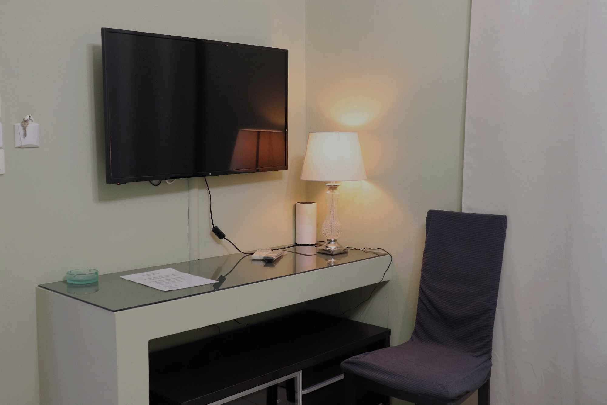 Quarto de hotel pequeno com televisão na parede, uma escrivaninha com luminária, uma cadeira e uma mesa de vidro.