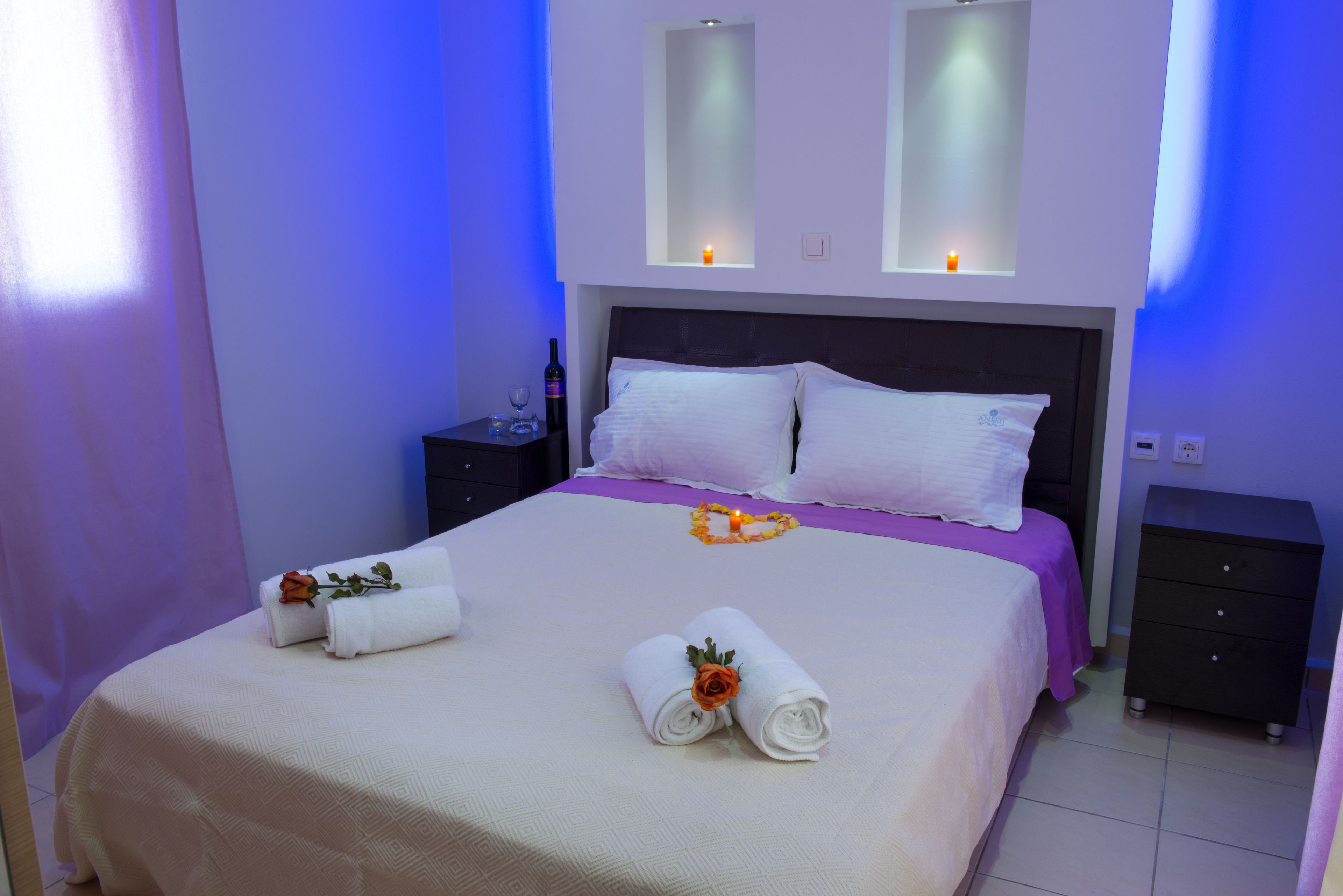 A neatly made bed with white linens and a rose, a bedside table with a bottle and glass, and a nightstand with a lamp.