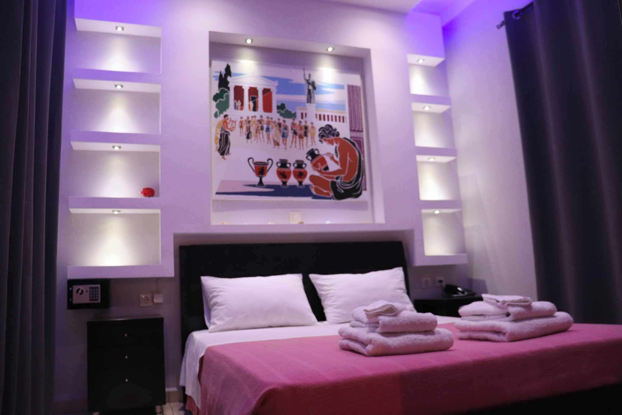 A hotel room with a bed, a nightstand, a wall-mounted control panel, and a painting above the bed.