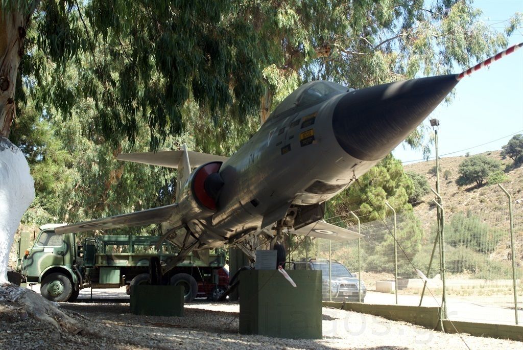 A military aircraft displayed outdoors, supported by a green vehicle and surrounded by trees.