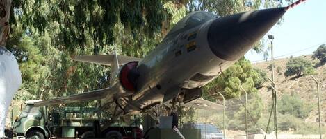 A military aircraft displayed outdoors, supported by a green vehicle and surrounded by trees.