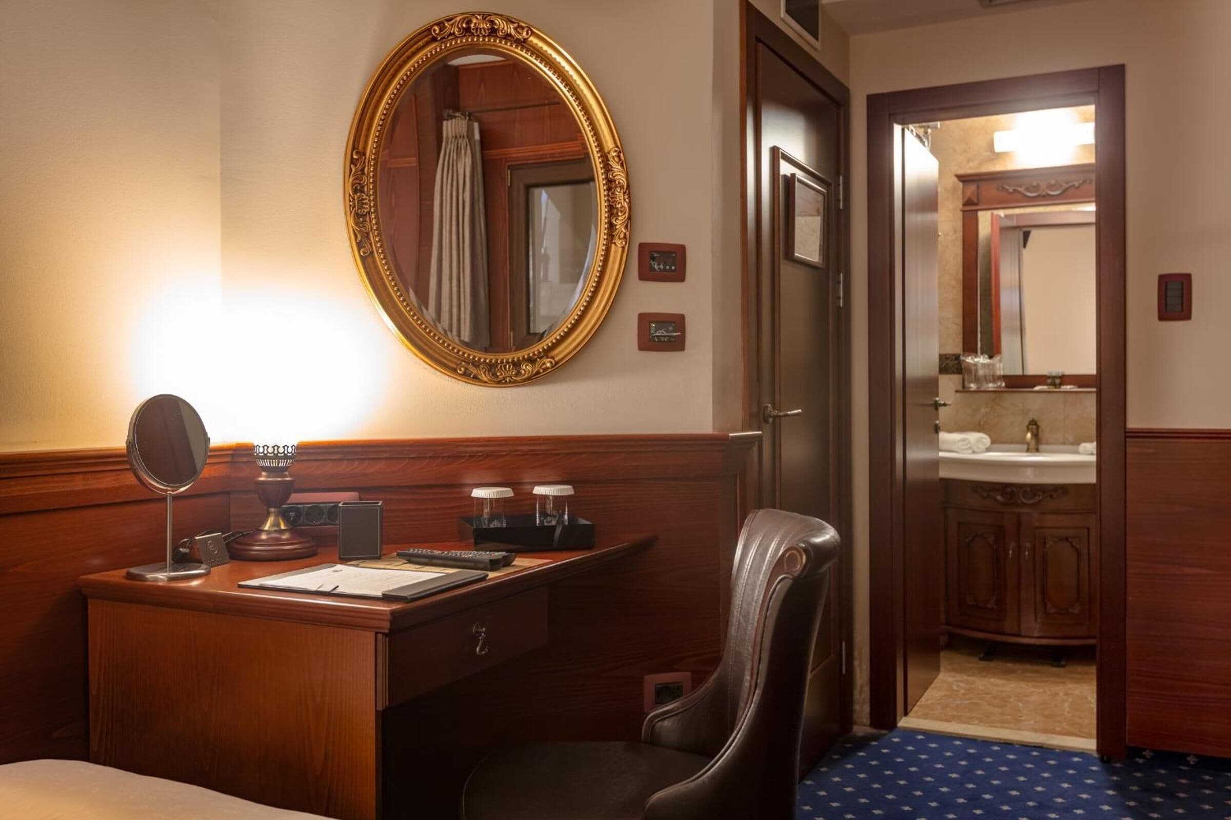 Habitación de hotel con escritorio de madera, una silla, un espejo grande y un baño visible a través de una puerta abierta.