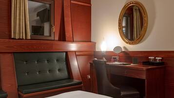 Habitación de hotel con cama, un banco tapizado en verde, un escritorio de madera con una silla, un espejo y una ventana con cortinas.