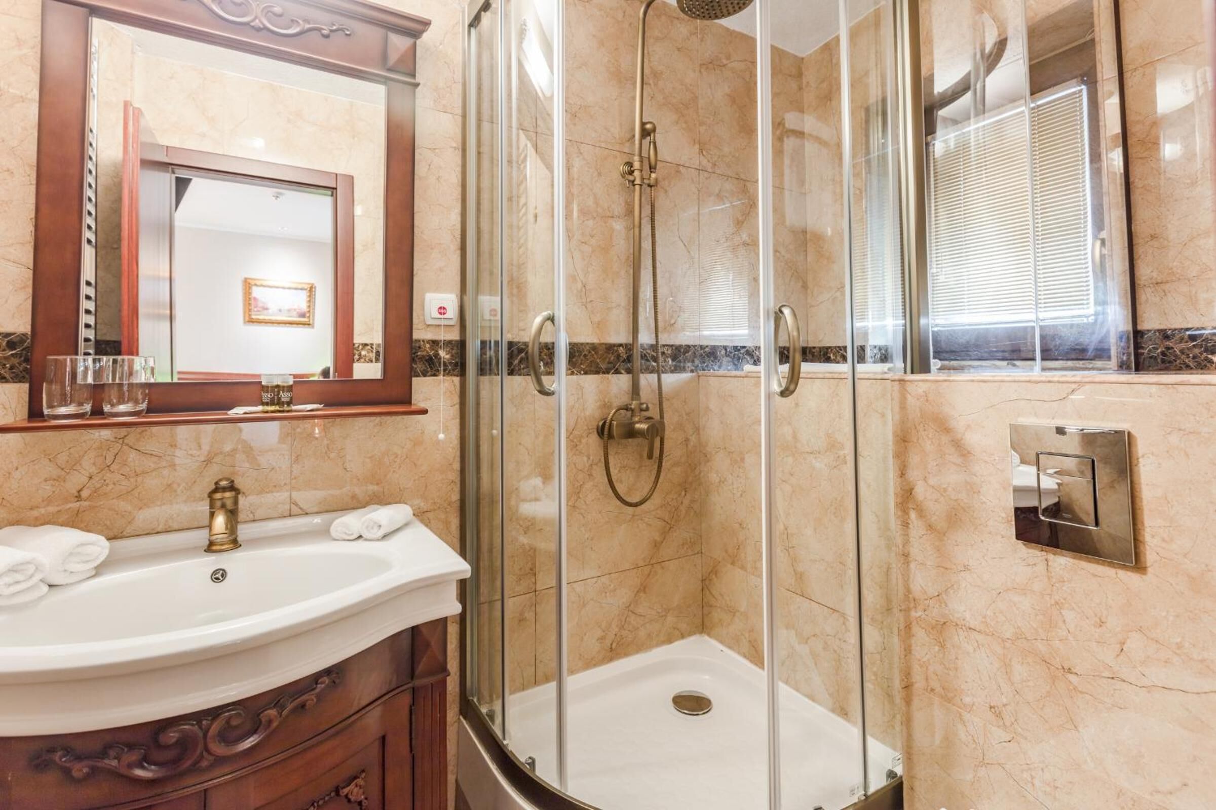Un baño con ducha envuelta en vidrio, pared con azulejos de mármol, lavamanos con grifo dorado y espejo con marco de madera.