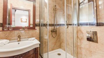 Un baño con ducha envuelta en vidrio, pared con azulejos de mármol, lavamanos con grifo dorado y espejo con marco de madera.