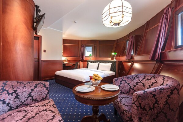 Une chambre d’hôtel avec un lit, deux fauteuils, une petite table avec des assiettes et des fruits, et un lustre.