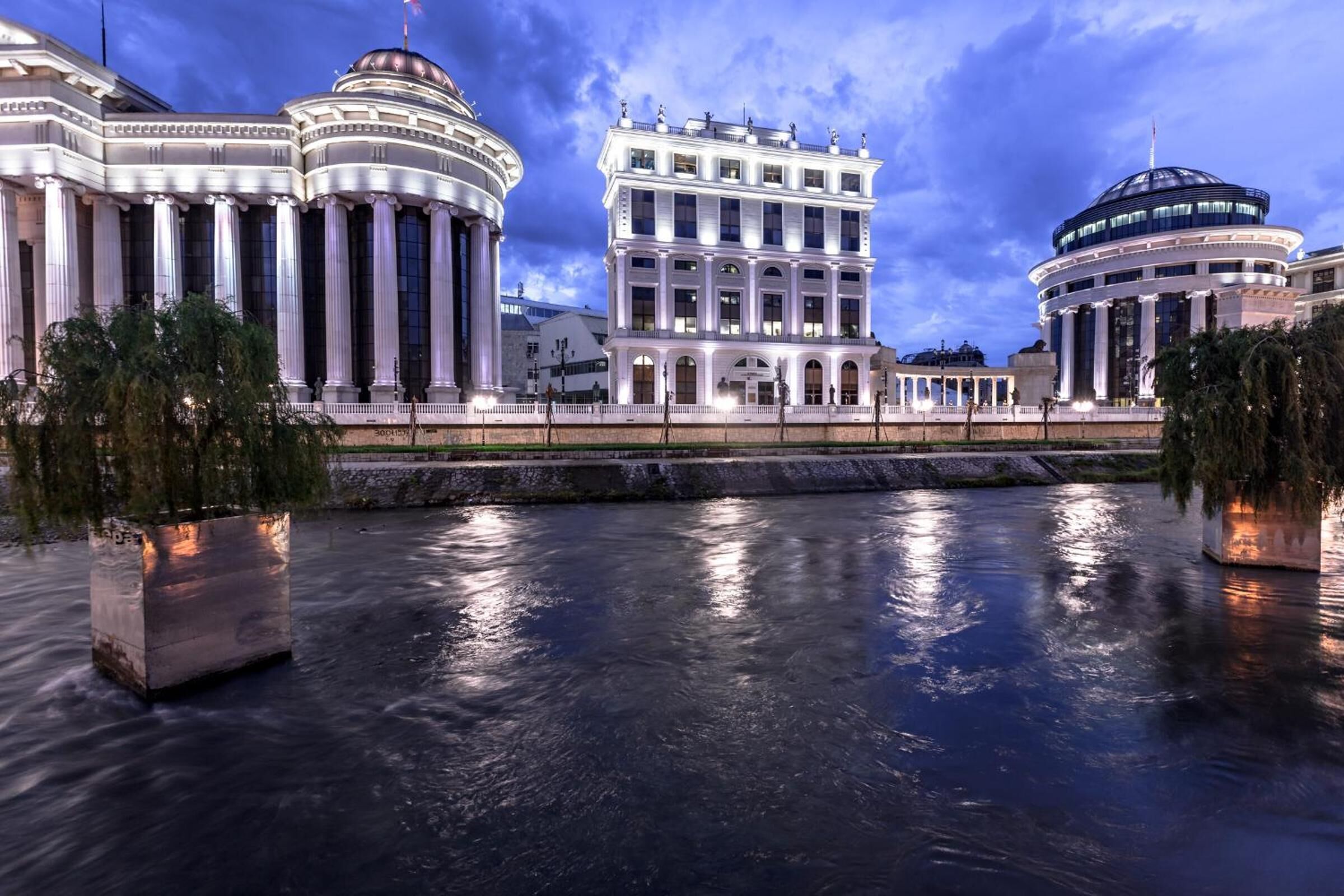 A grand building with classical architecture, featuring columns and domes, situated beside a calm river.
